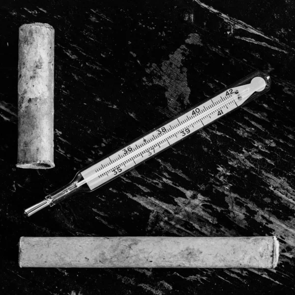 old medical mercury thermometer and paper case on black grunge wooden background close-up top view, black and white photo