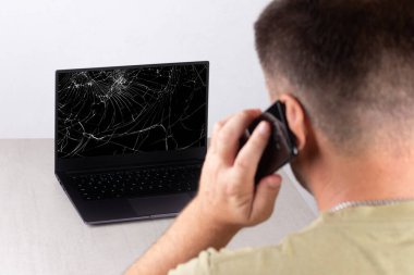 Bilgisayarın önünde cep telefonuyla konuşan genç bir adam kırık bir ekranla, teknik destek veya bilgisayar onarım servisiyle iletişime geçme konseptiyle,