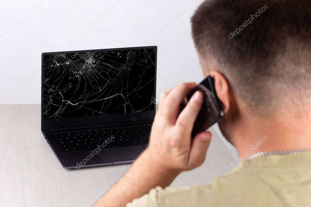 a young man talking on a mobile phone in front of a laptop with a broken, cracked screen, the concept of contacting technical support or computer repair service