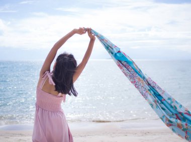 Pembe elbiseli ve şapkalı Asyalı kadın kumsalda, bulanık deniz görüntüsüyle dinleniyor. Tatillerde seyahat için.