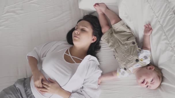 Молода стомлена мати спить біля плакального малюка в ліжку — стокове відео