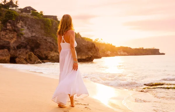 在日落的海滩上漫步在白色礼服的无忧无虑的妇女 — 图库照片