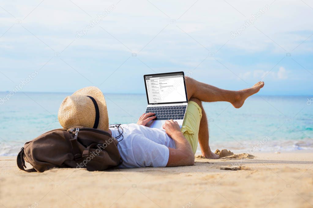 Man reading email on laptop while relaxing on beach