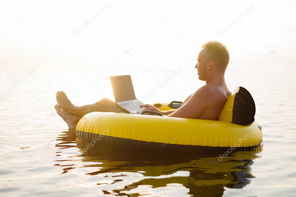 Businessman with a laptop on inflatable ring in the water at sunset