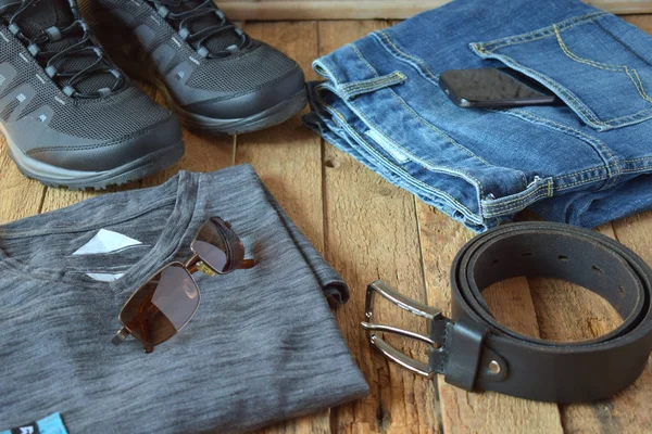 Men's casual outfits. Men shoes, clothing and accessories on wooden background - grey t-shirt, blue jeans, sneakers with eyeglasses, belt. Top view. Flat lay
