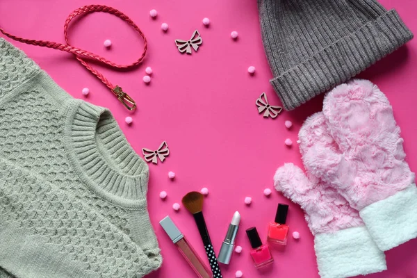 Knitted sweater, warm mittens, hat, belt, nail polishes, lipstick on a pink background. Winter and autumn set of warm, comfortable clothes and makeup cosmetics. Copy space. Top view.