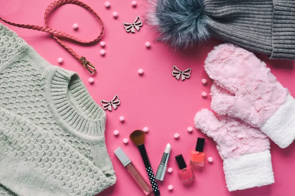 Knitted sweater, warm mittens, hat, belt, nail polishes, lipstick on a pink background. Winter and autumn set of warm, comfortable clothes and makeup cosmetics. Copy space. Top view.