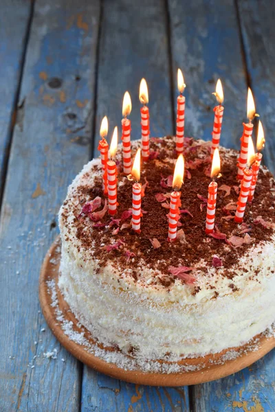 Delicious coconut layer cake - biscuit and cream on coconut cream, decorated with grated chocolate and candied rose petals. On the cake there are twelve red burning candles