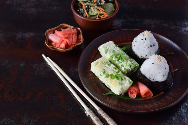 Mischung aus japanischem Essen - Reisbällchen onigiri, Omelette, eingelegter Ingwer, Sunomono Wakame Gurkensalat und Essstäbchen. asiatisches Frühstück oder Mittagessen Hintergrund — Stockfoto