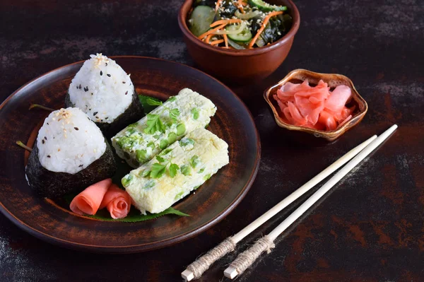 Mischung aus japanischem Essen - Reisbällchen onigiri, Omelette, eingelegter Ingwer, Sunomono Wakame Gurkensalat und Essstäbchen. asiatisches Frühstück oder Mittagessen Hintergrund — Stockfoto
