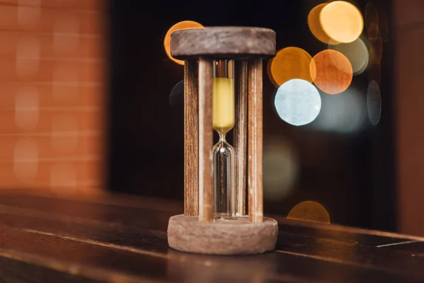 Vintage wooden hourglass against brick wall background and blurred lights. Time is running
