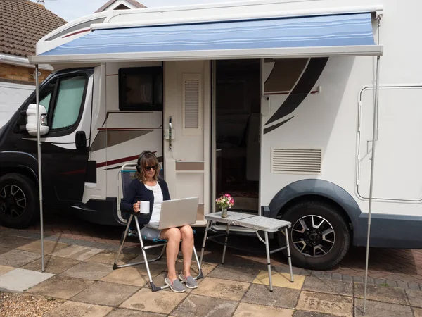 A lady motorhome owner is parked on her drive unable to travel due to Coronavirus lockdown.She has laptop headphones and mug sat on camping chair under awning of her recreational vehicle.Image