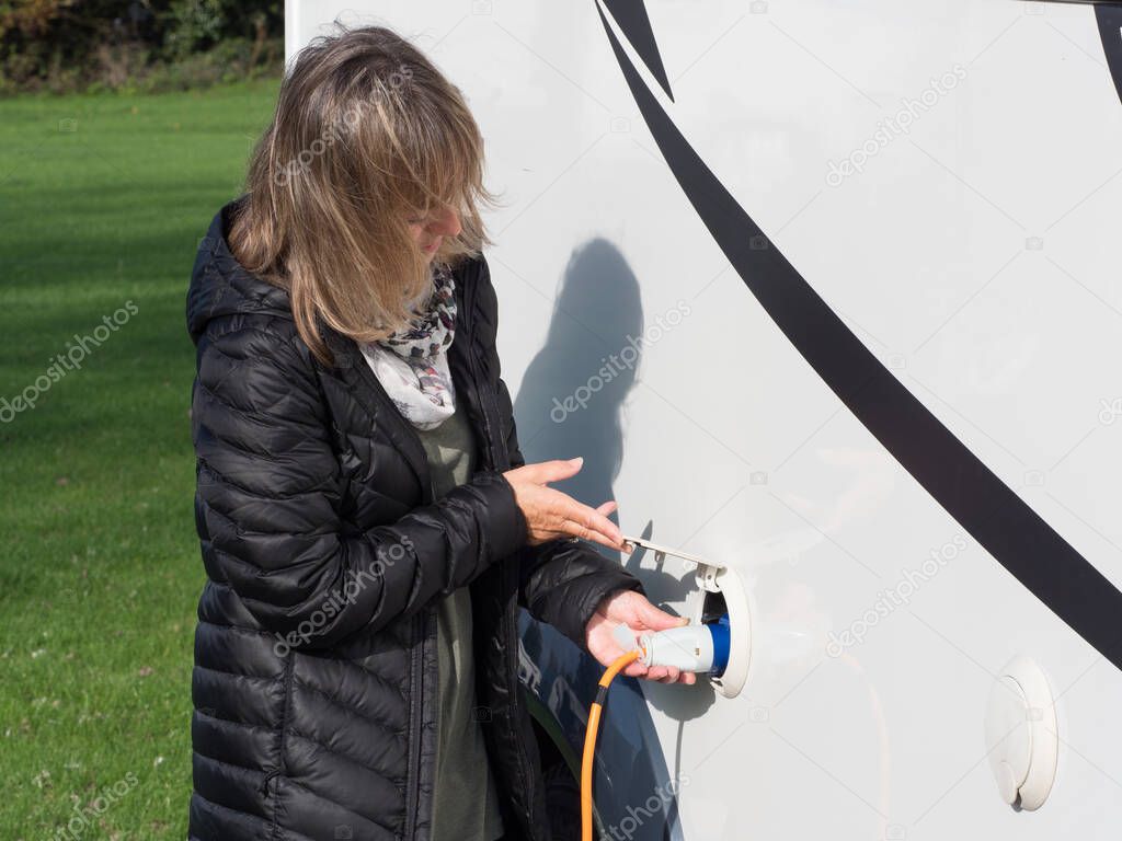 A lady lifts a cover flap and plugs in an orange electric hook up lead into the side of her motorhome to provide power to the camper van.Image