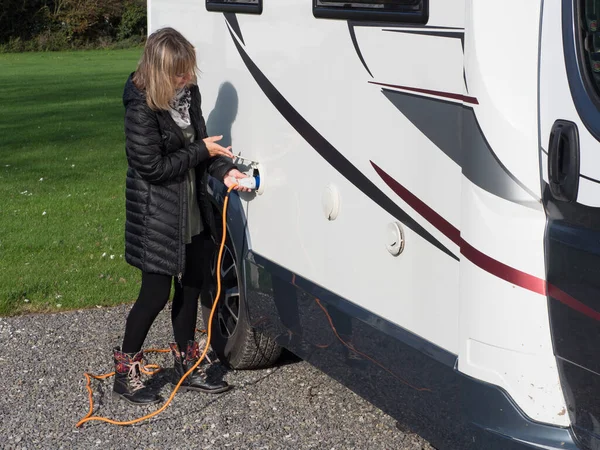 A lady standing lifts a flap and plugs in an orange electric hook up lead into the side of her motorhome to provide power to the recreational vehicle