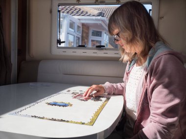 Karavanında oturan bir kadın pencere ışığı ile beyaz bir masada tamamladığı yapbozu aydınlatıyor..