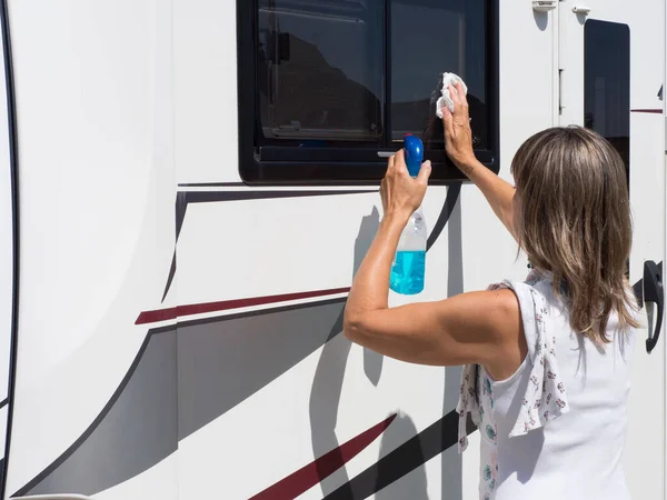 A lady motorhome owner cleans her windows.She has a spray gun with blue cleaning liquid in one hand and cleans the window with the other.Cloth over shoulder