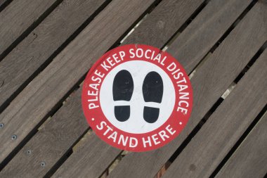 Tahta kaldırımdaki dairesel bir tabela 'lütfen sosyal mesafeyi koruyun' ve 'burada durun' diyor. Ayak izi sembolleri. Resim