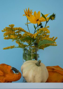 Balkabaklı vazoda çiçekler var. Çiçekler masada ve onların yanında balkabakları var.