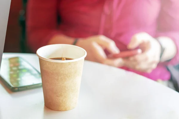 coffee in a recycled paper container with a girl checking her smart phone on the background
