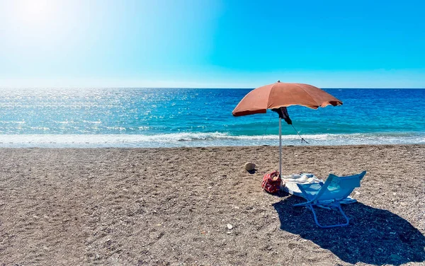 A deckchair under an umbrella facing the blue sea in a stony beach in Sicily, Italy. Summer holidays in Italian seaside resorts. Relax