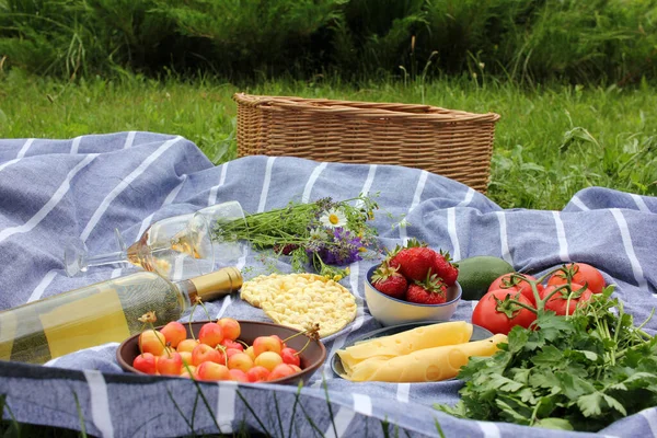 Summer picnic food. Bottle of white wine, glasses, strawberries, cherries in a bowl, fresh vegetables, tomatoes, parsley, avocado, rice crackers on a striped gray and white blanket. Top view