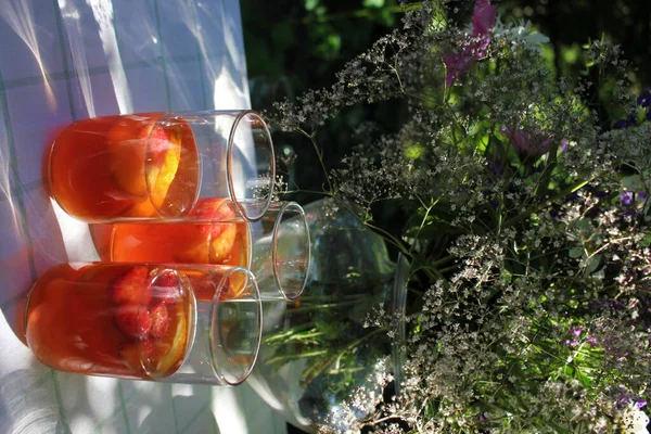 Glass ow white sangria and summer flowers on the cloth in the garden. White wine with fruits, oranges, strawberries and peaches in the glasses on the outdoor party