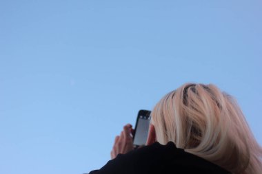 Pembe saçlı sarışın kız mavi gökyüzü arka planında uzanmış bir el telefonuyla akıllı bir telefon tutuyor. Arkadan bak. Selfie çeker, fotoğraf çeker, ya da bir ağ arar.