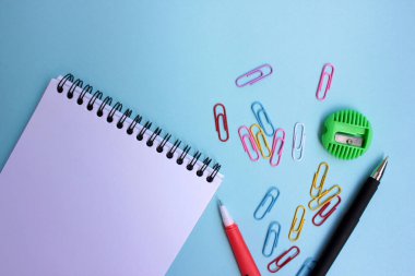 Mavi arka planda okul kırtasiyesi. Okul konseptine geri dönelim. Üst görünüm, boşluğu kopyala. Kağıt defteri aç, kırmızı kalem, silgi, yeşil kalem açacağı ve renkli ataçlar..