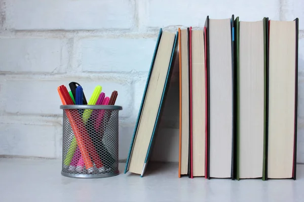 Stack of books and felt-tip pens on the table against white brick wall. Back to school background. Copy space