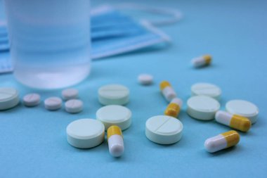 Beyaz haplar, sarı kapsüller, el dezenfektanı şişesi ve mavi arka planda tek kullanımlık tıbbi maske..