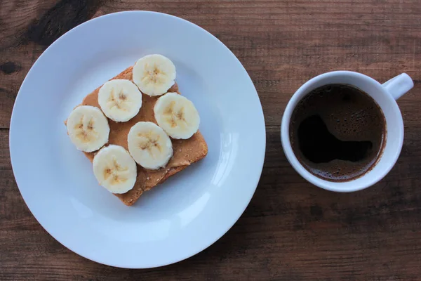 Cup of coffee and peanut butter banana toast on wooden background. Slices of whole wheat bran bread with peanut paste on a plate. Healthy breakfast