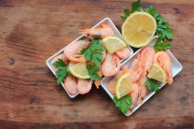 Maydanoz ve limonla servis edilen tabakta pişmiş pembe karides. Kabuklu Kuzey Atlantik karidesleri kırsal ahşap masadaki beyaz tabakalarda. Üst görünüm, alanı kopyala