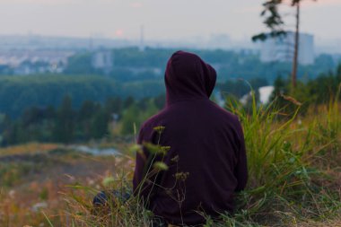 Sırtı çimenlerin üzerinde oturan ve yaz akşamları kırmızı ceketli şehre bakan bir adam.
