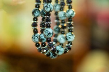 Yakın plan, odaktan uzak deniz kabuğu, artı mücevher, bilezik ya da kolye. Boncuklar birden fazla iplikte sıralanmış. Bu da nesneye katmanlı bir bakış veriyor.