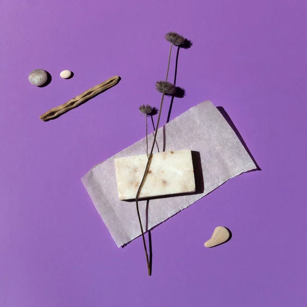 A bar of natural organic handmade cream-colored soap on a beautiful violet background surrounded by sea stones and dry herbs, close-up, top view. Abstraction, minimalism.