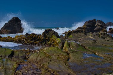 Öğle vakti, Sawarna Sahili 'nin batısındaki Java Endonezya' da bulunan bulutlu mavi gökyüzüne karşı büyük kayaların oluşturduğu doğal manzara dalgalara meydan okur gibi kümeler oluşturuyor.
