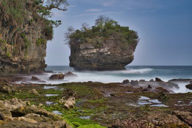 Öğle vakti, Sawarna Sahili 'nin batısındaki Java Endonezya' da bulunan bulutlu mavi gökyüzüne karşı büyük kayaların oluşturduğu doğal manzara dalgalara meydan okur gibi kümeler oluşturuyor.