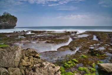 Öğle vakti, Sawarna Sahili 'nin batısındaki Java Endonezya' da bulunan bulutlu mavi gökyüzüne karşı büyük kayaların oluşturduğu doğal manzara dalgalara meydan okur gibi kümeler oluşturuyor.