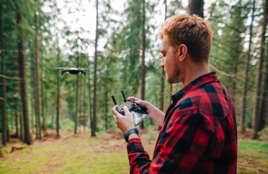 Yoğun bir ormanda, akıllı telefonla uzaktan kumandalı bir dronu, konsantre turist kontrol ediyor. Adam uzaktan kumandayla ormanda İHA uçuruyor. Pilotun ellerine odaklan..