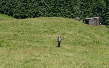 Erkek yürüyüşçü sırtında bir sırt çantasıyla bir dağ çayırında bir ahır ve ormanın arka planına doğru yürüyor. Dağlarda bir çayırda ve bir dağ köyünde yürüyüş yapmak