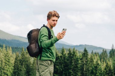 Sırt çantalı ve günlük giysiler içinde odaklanmış genç bir adam, dağ kozalaklı ormanı olan güzel bir manzaranın arka planına karşı akıllı telefon kullanıyor. Adam dağlarda internet kullanıyor.