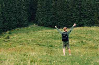 Bulanık bir fotoğraf. Ağaçlara odaklan. Yeşil kazaklı mutlu bir adamın arka görüntüsü. Bir dağı fethetti, zafer kazanarak ellerini kaldırdı.