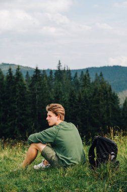 Oturmuş bir adamın dikey görüntüsü, sarışın, uzaklığa bakıyor, bir yaz dağı manzarası. Turist bir çayırda dinleniyor, başka tarafa bakıyor..