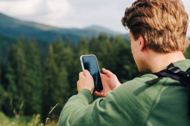 Telefonla seyahat ettiği güzergahına bakan bir adamın arka görüntüsü, dinlenmek için çimlerin üzerine oturdu. Akıllı telefondan internet kullanan bir turistin yakın fotoğrafını çek.