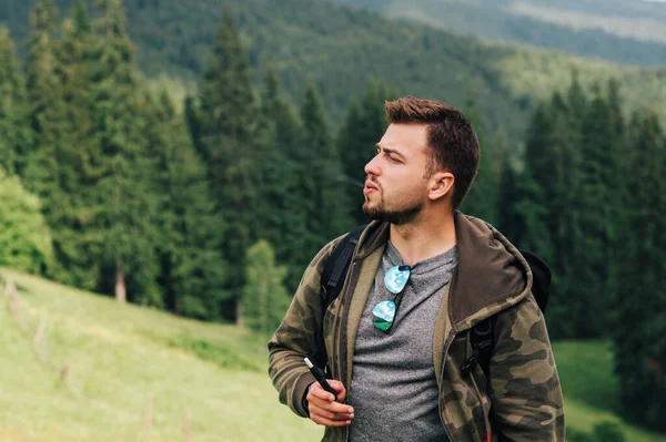 Portrait of a bearded man smokes a device for evaporating tobacco in mountains on the background of a beautiful landscape with a coniferous forest.Tourist smokes an electron cigarette on mountain hike