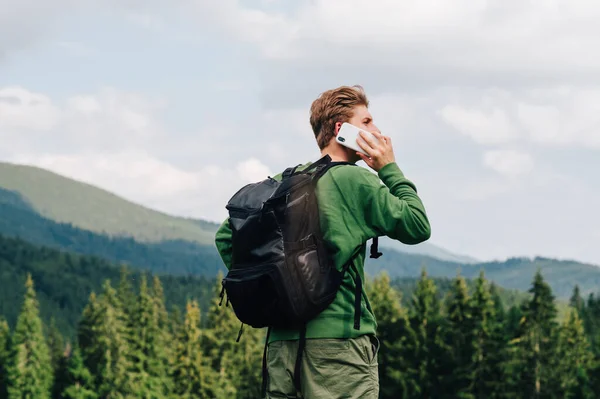 Guy in casual clothes stands on top of a mountain on a hike and calls on the phone, carries a backpack. Young male tourist talking on the phone in the mountains and looking away