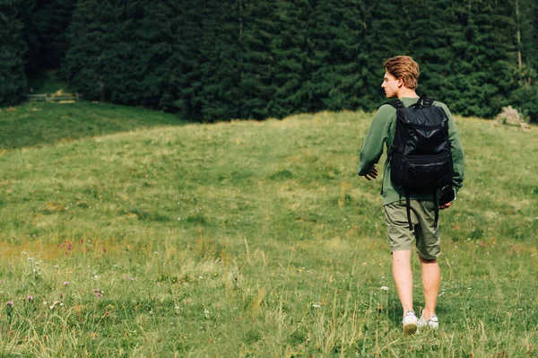 Young man, tourist, walking on a field in the mountains with a black backpack. Young hiker on a hike walks on a mountain meadow against the backdrop of a coniferous forest and looks away