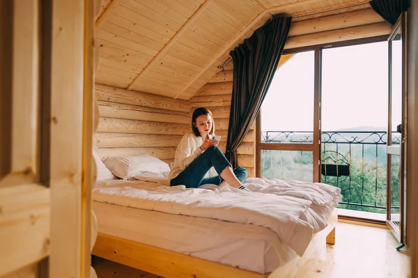 Beautiful woman in casual clothes sits on a bed in the apartment of a wooden country house and uses a smartphone with a serious face. Leisure in a country house.