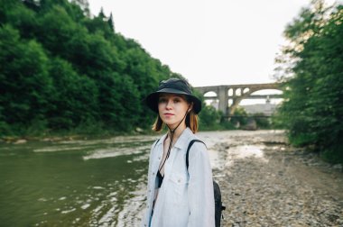 Bir dağ nehri ve köprünün arka planında duran şapkalı çekici bayan yürüyüşçü ciddi bir yüzle kameraya bakıyor. Dağ nehri ve orman boyunca güzel buz yürüyüşleri