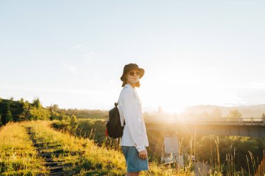 Günlük kıyafetler, şapka ve güneş gözlüğü takan çekici bir kız eski bir demiryolu köprüsünde duruyor ve güzel bir günbatımının arka planındaki kameraya bakıyor. Turizm konsepti.